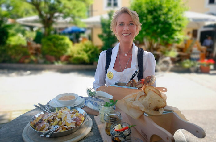 Landgasthäuser. Postwirtin Elisabeth Walch präsentiert Gerichte an einem Holztisch vor ihrem Gasthaus. Bild: Sender / BR / Bewegte Zeiten Filmproduktion GmbH / Carsten Frank